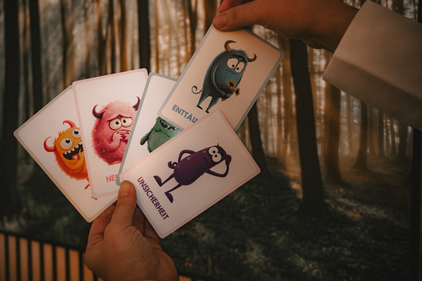Hände halten farbige Monsterkarten vor einer unscharfen Waldkulisse.